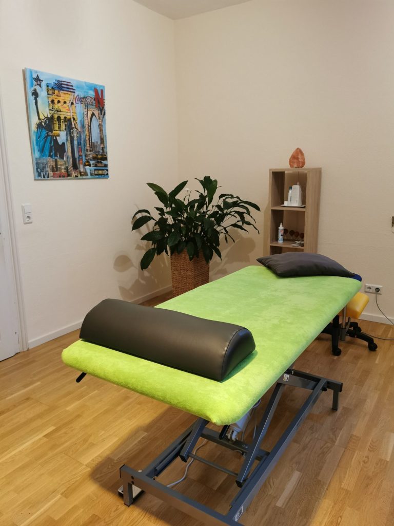 Behandlungsraum Physiotherapie mit Behandlungsliege, Gymnastikball und Bodenmatten. In einem Regal stehen Lotionen und Behandlungsmaterialien; darauf eine Pflanze. Der Raum ist in warmen Farben gehalten.