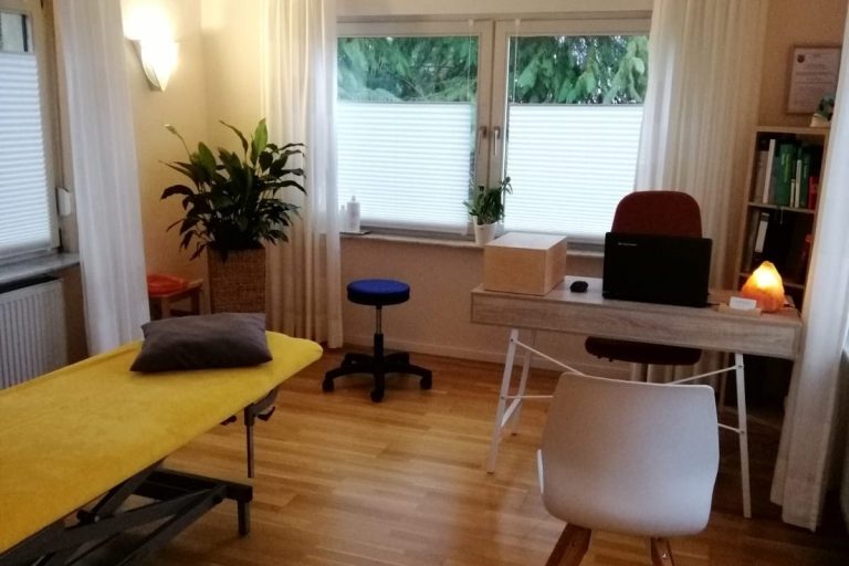 Behandlungsraum Osteopathie mit Behandlungsliege, Schreibtisch, Stühlen, einer großen Pflanze und einem Bücherregal. Der Raum ist in warmen Farben gehalten.