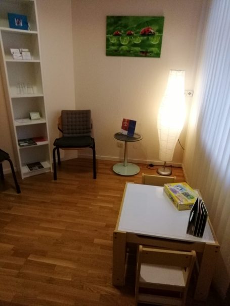 Das Wartezimmer mit Stühlen, einem Regal und einem Tisch mit Informationsmaterial; Kinderecke, Stehlampe und ein Wandbild mit Marienkäfern.