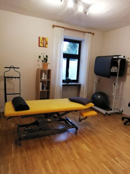 Behandlungsraum Physiotherapie mit Behandlungsliege, Gymnastikball und Bodenmatten; dazu ein Regal mit Lotionen und Behandlungsutensilien.