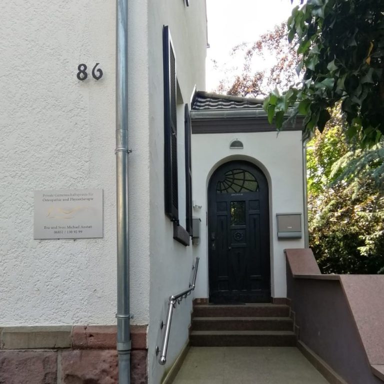 Hauseingang der Praxis mit wenigen Treppenstufen, die zu einer oben gerundeten Haustür führen, daneben die Hausnummer 86 und das Praxisschild.