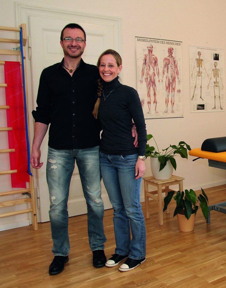 Sven Michael und Eva Anstatt auf einer kleinen Treppe vor dem Praxiseingang und dem Praxis-Schild.